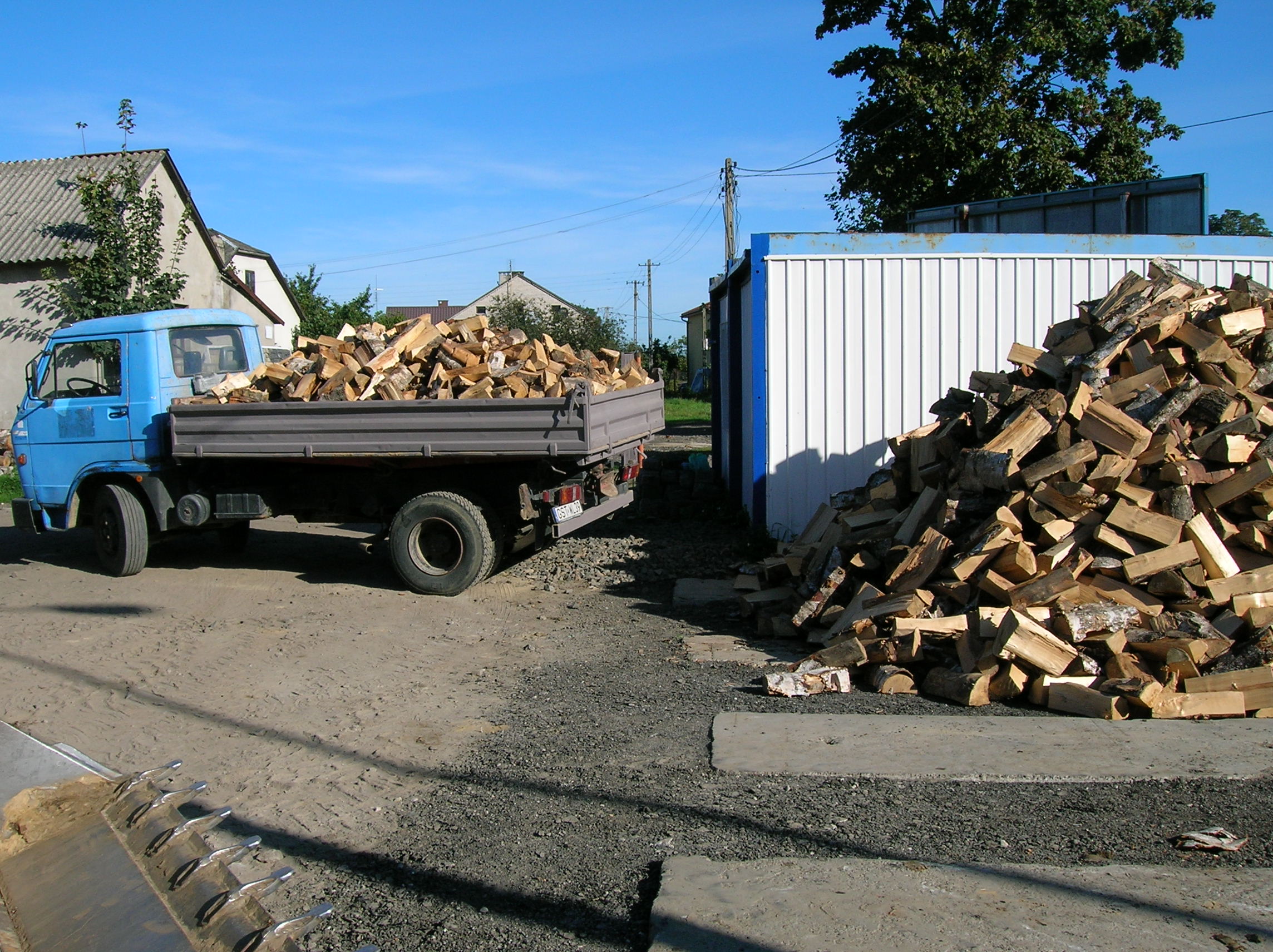 Niebieska ciężarówka z drewnem kominkowym obok białego kontenera, na ziemi usypana sterta pociętego drewna, w tle budynki mieszkalne pod błękitnym niebem.